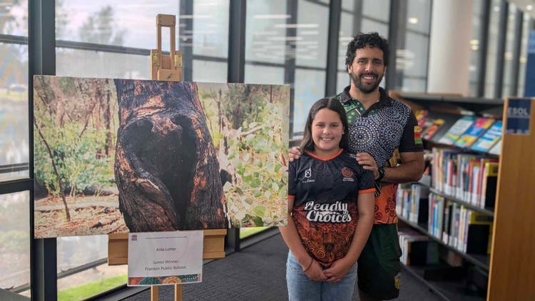 Student Aida Lotter standing beside her father, smiling next to a framed piece of her artwork on display.